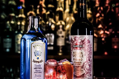 Ginebra y vermut junto a dos cócteles con el botellero iluminado al fondo